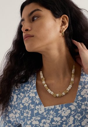 Perlekæde med cremefarvede og gule cirkulære og rektangulære perler. Båret med en blå topp med blomsterprint. Håret er mørkt og bølget.