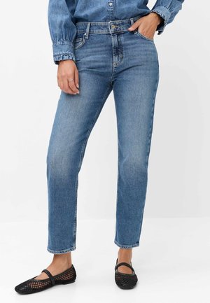 Kvinde iført blå denim-jeans, en blå denim-skjorte og sorte mesh-flade sko, stående med den ene hånd i lommen og den anden hånd afslappet.