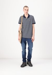 Marinblå och vit randig polo, denimjeans, svarta sneakers och en beige axelväska. Modellen står framför en neutral bakgrund.