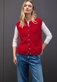 Röd stickad ärmlös kofta med knappframsida, lager-på-lager över en vit långärmad skjorta; kombinerad med blå denimjeans.