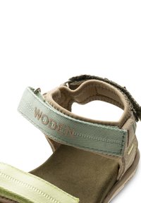 Ruskea ja vihreä tekstiilisandaali, jossa on säädettävät hihnat, mokkanahkainen pohja ja vaaleanvihreät yksityiskohdat. Hihnassa on "WODEN"-logo.