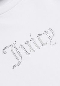 Vit T-shirt med ordet "Juice" prytt med silverfärgade rhinestones, som visar en elegant kursiv design och slät tygtextur.