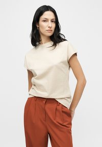 Μπεζ κοντομάνικο μπλουζάκι από μαλακό ύφασμα, συνδυασμένο με ψηλόμεσα παντελόνια σε σκουρόχρωμο σκουριασμένο χρώμα με πιέτες και εφαρμοστό κόψιμο.