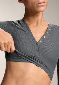 Szürke bordázott felső V nyakkivágással és elülső gombokkal, testhez simuló szabással és puha textúrával, részben felhúzva, hogy felfedje az alsó réteget.