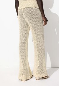 Pantalons évasés en tricot beige clair avec une texture à mailles ouvertes. Présente une taille et un ourlet festonnés, mettant en valeur le design aérien.