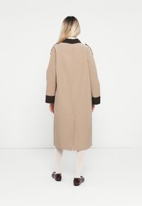 Frau mit langen blonden Haaren, die einen beigen langen Mantel mit dunklen Bündchen und Kragen trägt, weiße Strumpfhosen und dunkle glänzende Ballerinas, steht mit dem Rücken zugewandt.