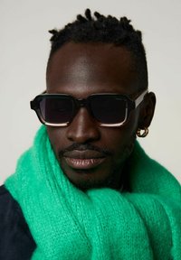 Svarta solglasögon med en bred rektangulär ram, klar nederkant och mörka linser, i kombination med en fluffig ljusgrön halsduk.