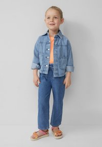 Denimjacke mit Knopfleiste, hellblaue Farbe, mit zwei Fronttaschen. Kombiniert mit einem orangefarbenen Oberteil, blauen Jeans und orangefarbenen Sandalen.