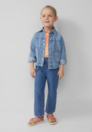 Denim jack met knopen aan de voorkant, lichtblauwe kleur, met twee voorzakken. Gecombineerd met een oranje top, blauwe spijkerbroeken en oranje sandalen.