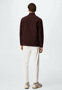 Giacca bordeaux scuro con colletto, abbinata a pantaloni bianchi. Il tessuto sembra testurizzato e le scarpe sono di un beige chiaro con una finitura liscia.
