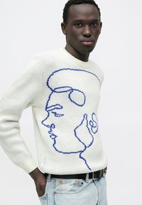 Witte gebreide trui met een blauwe lijntekening van een gezicht. Geribbelde ronde hals en manchetten, gedragen met lichtblauwe denim en een zwarte riem.