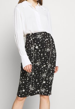 Kobieta w białej bluzce z długim rękawem i czarnej ołówkowej spódnicy do kolan z białymi wzorami w kształcie gwiazd oraz pionowymi szwami.