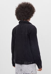 Svart denimjacka med shearlingkrage, med stickade detaljer, en ryggparti och knappar vid nederkanten. Visas från baksidan.