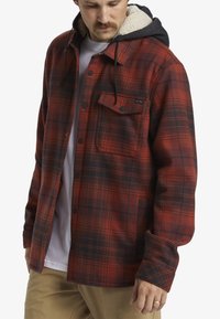 Red and black plaid flannel shirt with a soft, beige sherpa-lined hood. Features snap buttons and a front pocket.
