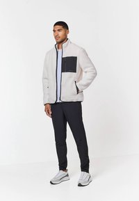 Giacca in pile crema con tasca nera, zip frontale e collo alto; abbinata a pantaloni neri e sneakers grigie; tessuto strutturato.