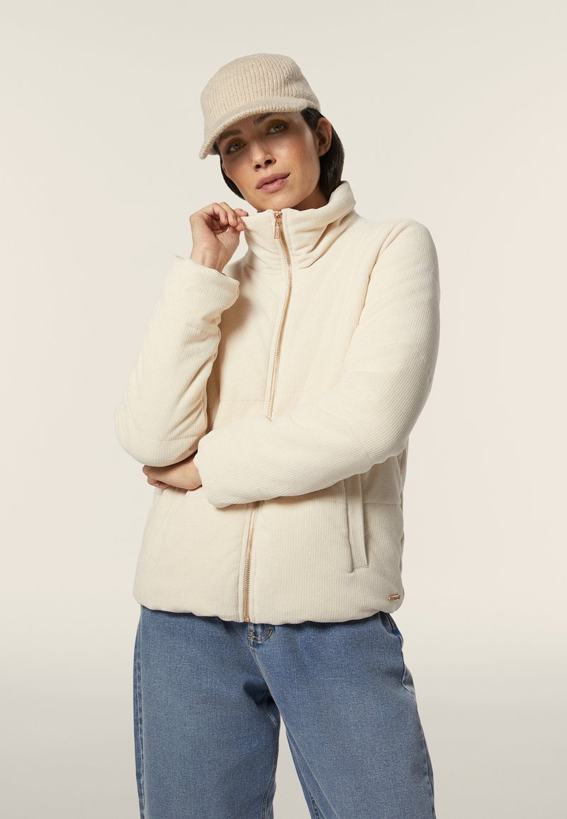 Chaqueta cremosa con cremallera y collar alto, textura acanalada y bolsillos laterales. Combinada con unos vaqueros azules claros y una gorra a juego.