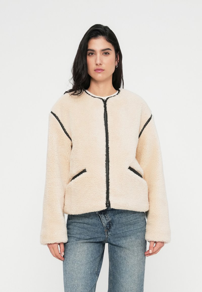 Ζακέτα φλις σε κρεμ απόχρωση με μαύρο φερμουάρ και διακοσμητικές πινελιές στις τσέπες. Διαθέτει στρογγυλή λαιμόκοψη και άνετη γραμμή με μακριά μανίκια. Μαλακή υφή.