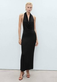 Vestido negro con cuello halter, cuerpo drapeado, silueta ajustada y diseño de largo hasta el suelo. Combinado con sandalias rojas de tiras y pendientes grandes.