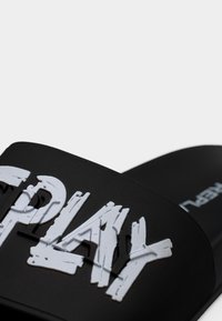 Μαύρη σαγιονάρα με λουρί με υφή και ακανόνιστα λευκά γράμματα που σχηματίζουν τη λέξη "PLAY." Μαλακό καουτσούκ με λείο φινίρισμα.