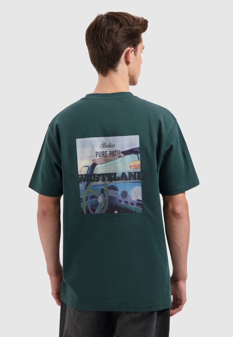 Grünes Baumwoll-T-Shirt mit einem rechteckigen Grafikdruck auf dem Rücken, der eine farbenfrohe Illustration eines Auto-Innenraums und Text in kontrastierenden Farben zeigt.