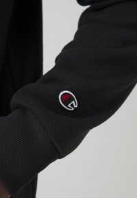 Czarna bluza z teksturowanej tkaniny, z prążkowanym mankietem i małą, czerwoną i białą łatką z logo na rękawie.