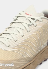 Botte de sport beige avec un dessus texturé, accents ondulés blancs, col rembourré et lacets gris. Elle possède une semelle en caoutchouc épaisse et un détail de logo.