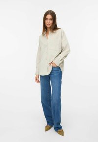 Light beige button-up shirt with a single chest pocket, paired with blue straight-leg jeans and pointed beige shoes, set against a plain background.