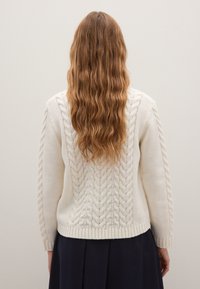 Cream-colored cable-knit sweater featuring textured patterns, long sleeves, and a ribbed hem. Displayed from the back with soft wavy hair visible.