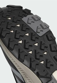 Zbliżenie na czarno-szarą, wytrzymałą podeszwę buta z wypukłymi, geometrycznymi wzorami bieżnika i trójkątnymi wycięciami.