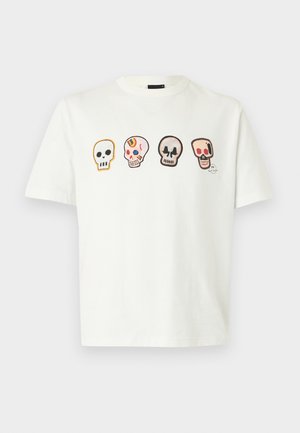 Jeune homme aux cheveux bouclés portant un t-shirt blanc avec quatre crânes colorés, un pantalon noir avec un cordon bleu, et des baskets blanches.