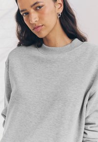 Pilka megztinio su tekstūruotu apvaliu iškirptu kaklu ir minkštu paviršiumi. Moteris su banguotais plaukais dėvi apskritus auskarus, žvelgianti šiek tiek link kameros.