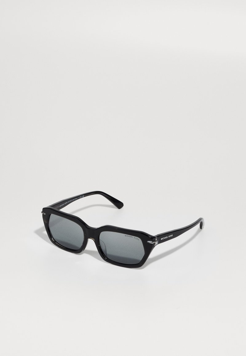 Melnas saulesbrilles ar taisnstūra formu, pelēkām tonišķēm lēcām un maziem metāla akcentiem. Zīmola nosaukums "Michael Kors" ir redzams uz kājas.