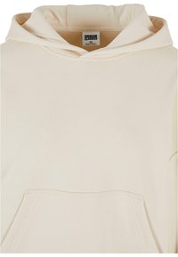 Haut à capuche crème clair en tissu doux, avec une grande capuche et une poche avant. Le design est minimaliste avec des lignes épurées et sans motifs.