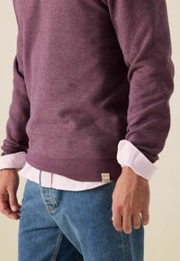 Maroon katoenen sweatshirt over een wit en roze gestreept shirt, met opgerolde mouwen. Blauwe denim jeans met zichtbare patchlabel.