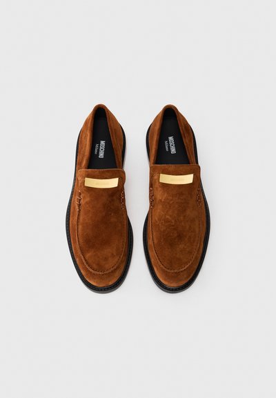 Sapatos deslizantes em camurça castanha com sola preta plana, apresentando um detalhe metálico dourado na parte superior e pormenores de costura suave.