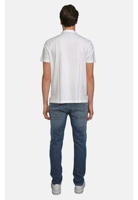Polo bianco con maniche corte, taglio dritto, indossato con jeans blu e sneaker bianche, visto da dietro. Design semplice, senza motivi.