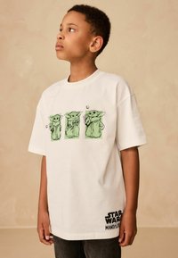Tricou alb din bumbac, având trei reprezentări grafice verzi ale unui personaj cu urechi mari, însoțite de textul negru "Star Wars Mandalorian".