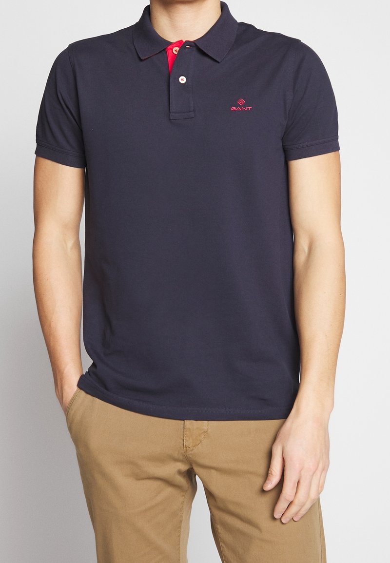 Polo shirt bleu marine en coton, avec un col rouge et une patte de boutonnage à trois boutons, arborant le logo GANT brodé en rouge sur la poitrine gauche.