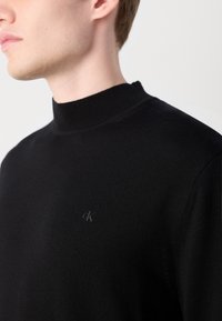 Črna pletena pulover z visokim ovratnikom ima rebrast ovratnik in malo izvezeno logo na prsih, kar zagotavlja gladko teksturo in prilagojen dizajn.