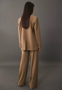 Femme aux cheveux longs et raides portant un blazer ample de couleur beige et un pantalon à jambes larges, debout face à un fond gris uni.