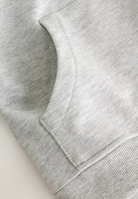 Primo piano di un tessuto di felpa grigio chiaro che mostra una tasca frontale con una texture liscia e dettagli a coste sui polsini. Leggero motivo melangiato visibile.