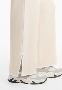 Pantalons large beige clair avec fentes sur les côtés, dotés d'une texture douce et de bandes blanches contrastantes, accompagnés de baskets grises.