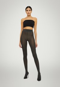 Svarta, genomskinliga strumpbyxor med hög midja, i kombination med en svart axelbandslös topp. Modellen har på sig spetsiga svarta klackar. Slät textur, åtsittande design.