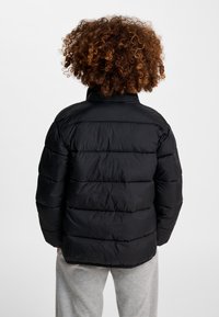 Person med krøllet hår iført en sort dunjakke og lysegrå bukser, vist bagfra mod en ensfarvet baggrund.