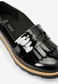 Chaussure en cuir verni noir avec un design plissé à l'avant, bout rond et semelle marron contrastante avec des accents de couture blanche.