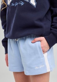 Pantaloni corti azzurri chiari con vita regolabile e tasche laterali, decorati con accenti bianchi e la scritta "New York" ricamata vicino all'orlo.