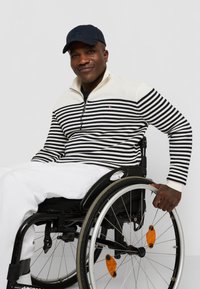 Gestreepte pullover in marineblauw en crème, met een halve rits en geribde boorden. Model in een rolstoel, met de hand op het wiel. Witte broek.