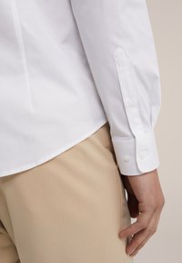 Witte langemouwen shirt met een gladde textuur, met knoopmanchetten en een puntige kraag, gecombineerd met beige broeken. Eenvoudig ontwerp met focus op het materiaal.