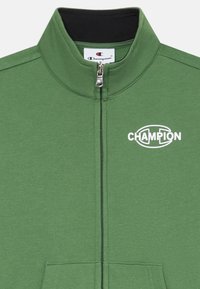 Chaqueta verde con cremallera y cuello plano, que presenta un logo blanco de "CHAMPION" en el lado izquierdo del pecho y bolsillos tipo canguro en la parte delantera, fabricada en tejido suave.