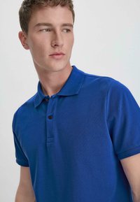Royal blue polo shirt made of textured fabric, featuring a collar, two brown buttons, and short sleeves. Sophisticated sporty design.
